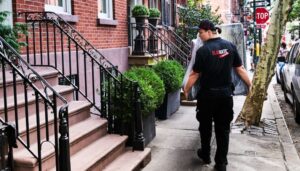 Man in a black shirt with the text "pilates" walking past brownstone steps on a city sidewalk.