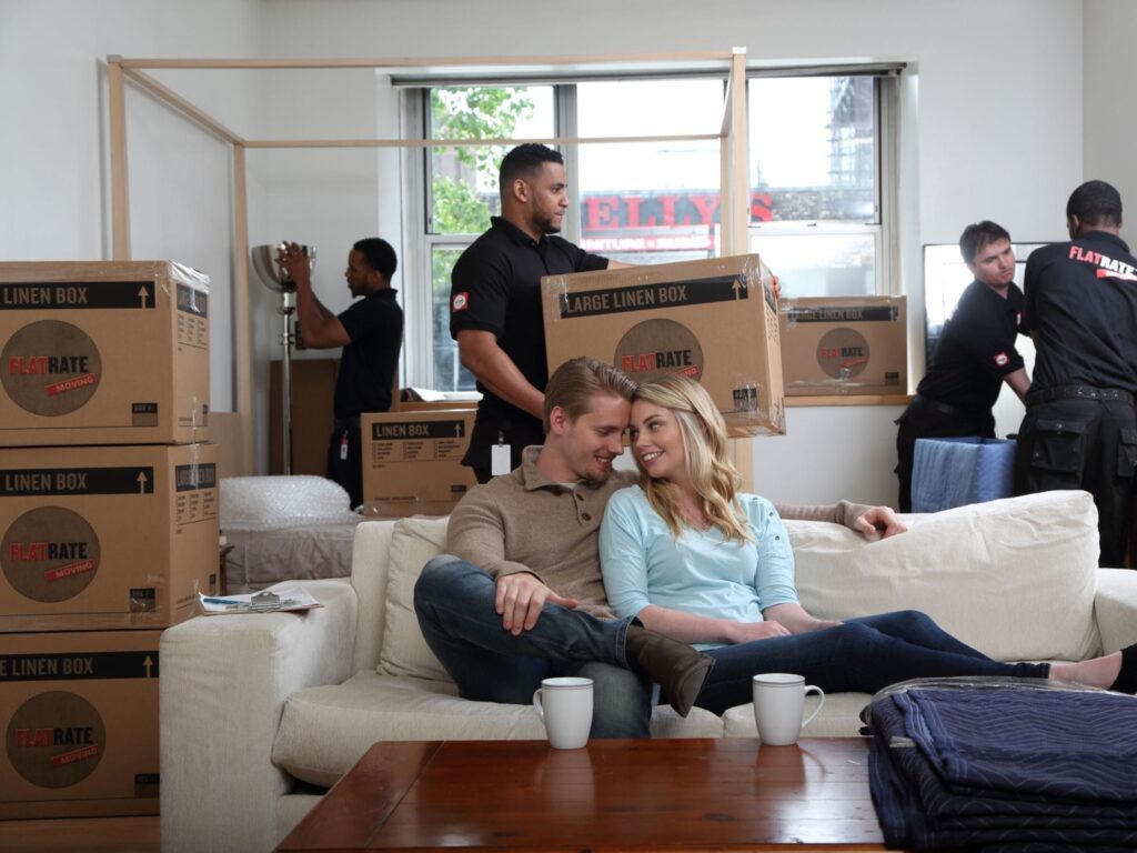 A couple sitting comfortably on a couch while movers pack up their belongings in the background.