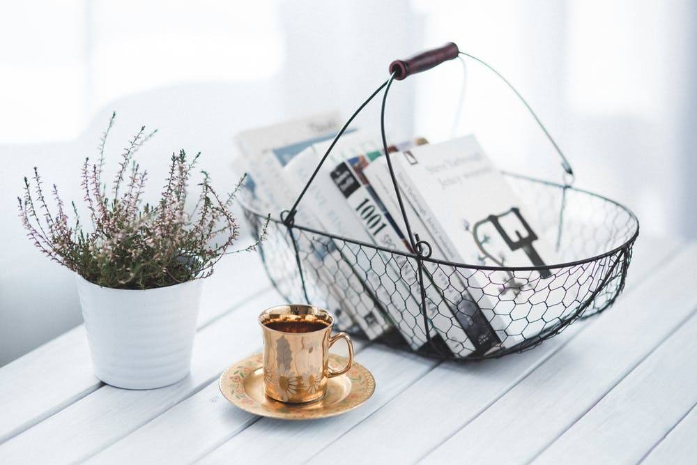 A serene setting with a vintage gold cup on a saucer, a basket of newspapers, and a potted plant on a white wooden table.