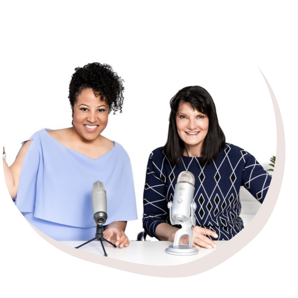 Two women smiling and sitting at a table with microphones, possibly recording a podcast.